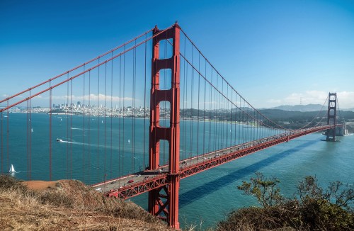 Golden Gate Bridge spanning a bay, with sailboats and a city skyline in the background under a clear blue sky.