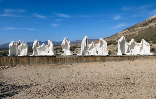 A series of white ghostly sculptures on a desert landscape, set against a clear blue sky and distant mountains.