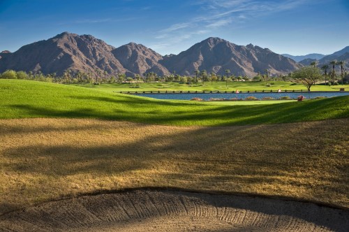 Lush green golf course with a pond and mountains in the background under a clear blue sky.