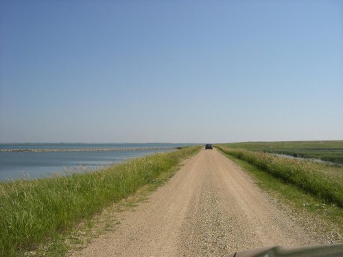 A gravel road stretches alongside a calm body of water, bordered by green grass under a clear blue sky.