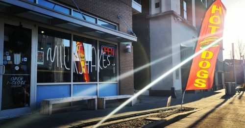 A storefront with a sign reading "Milkshakes" and a bright "Hot Dogs" flag outside, illuminated by sunlight.