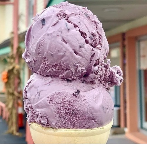 A close-up of a double scoop of purple ice cream in a cone, set against a blurred background of a street.