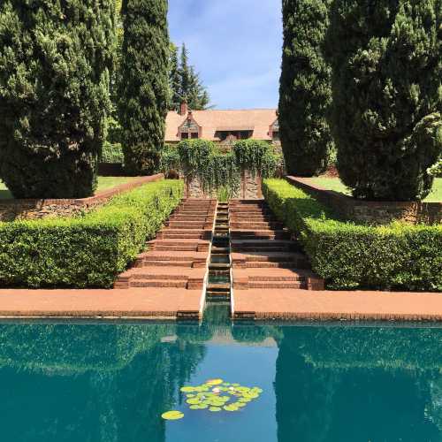 A serene garden with neatly trimmed hedges, a reflecting pool, and a grand house in the background under a clear blue sky.