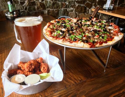 A large pizza topped with meats and vegetables, alongside a basket of chicken wings and a pitcher of beer.