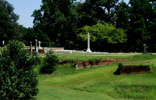 A serene cemetery scene with gravestones and a tall white monument surrounded by lush green trees and grass.