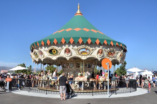 A colorful carousel with a green and orange canopy, surrounded by people enjoying a sunny day at a fair.