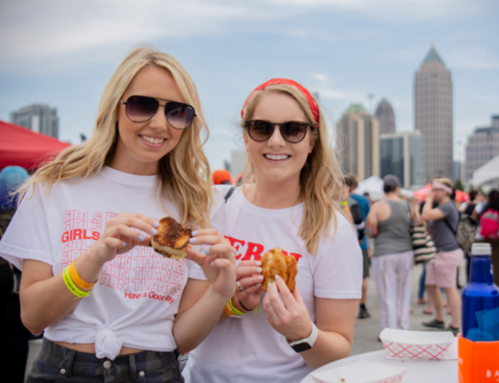 Two smiling women in sunglasses hold food at an outdoor event, with a crowd and city skyline in the background.
