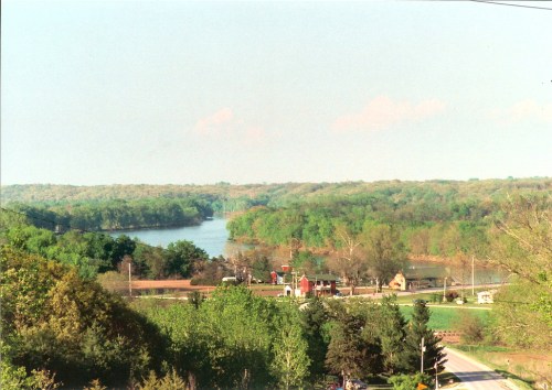 A scenic view of a river surrounded by lush green trees and a small rural settlement in the foreground.