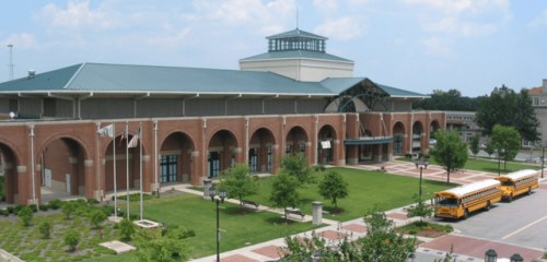 A large brick building with a green roof, surrounded by trees and school buses parked in front.