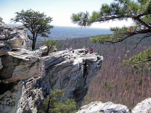 A group of people sitting on a rocky cliff edge, surrounded by trees and a vast landscape in the background.