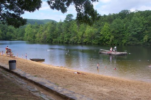 A sandy beach by a calm lake, with people swimming and a dock in the background surrounded by lush green trees.