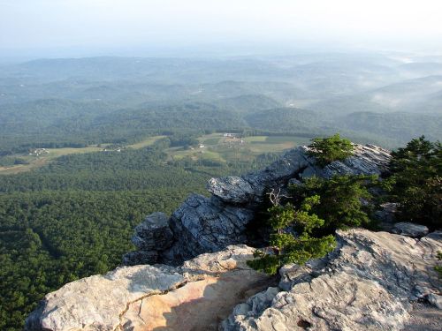 A panoramic view from a rocky mountain peak overlooking lush green valleys and distant hills under a clear sky.