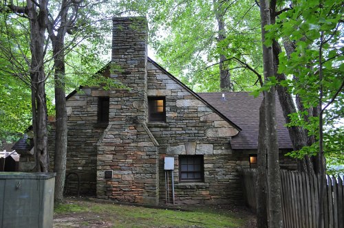 A rustic stone house surrounded by trees, featuring a chimney and a sloped roof, set in a natural landscape.