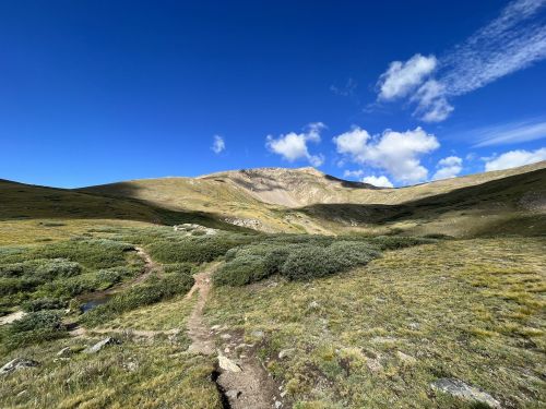 A scenic mountain landscape with a clear blue sky, grassy terrain, and a winding dirt path leading towards the peak.