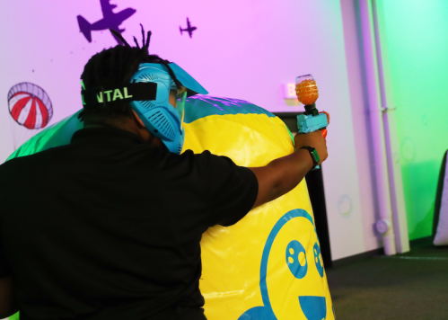 A person wearing a blue mask aims a toy gun at a colorful inflatable target in a vibrant indoor setting.
