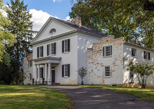 A historic white house with columns, surrounded by trees and a grassy area, showing signs of age on the exterior.
