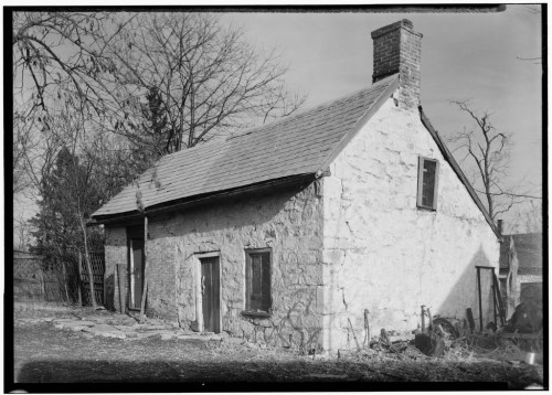 A weathered stone cottage with a chimney, surrounded by bare trees and a grassy area.