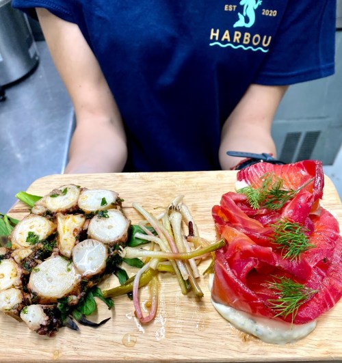 A wooden platter featuring grilled octopus, pickled vegetables, and thinly sliced fish garnished with dill.