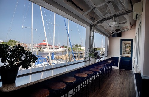 A waterfront restaurant interior with a bar, stools, and large windows overlooking boats and a marina.