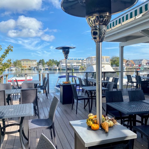 Outdoor dining area by a waterfront, featuring tables, heaters, and autumn decorations with pumpkins.