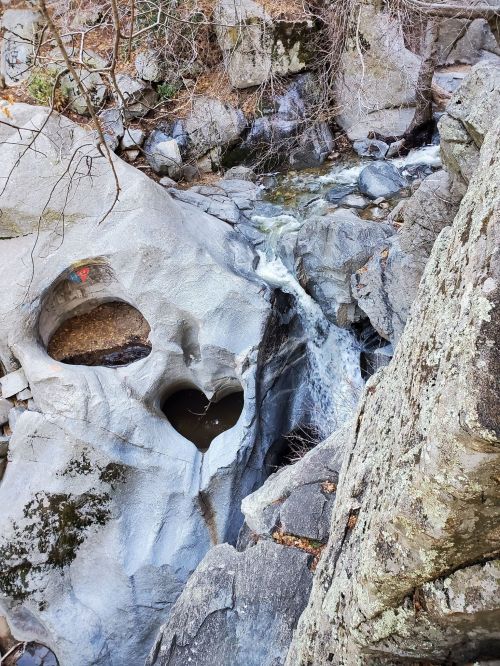 A heart-shaped hole in a rocky cliff beside a small waterfall and flowing stream in a natural setting.