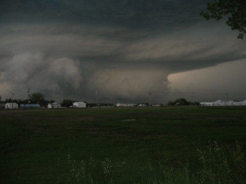 Dark, ominous storm clouds loom over a rural landscape with scattered buildings and a grassy field.