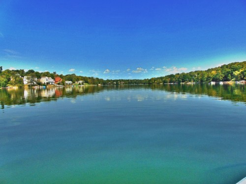 A serene lake scene with clear blue water reflecting trees and houses under a bright blue sky.