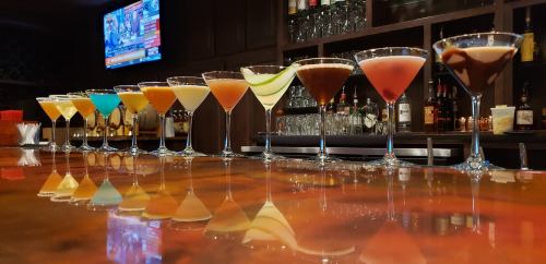A row of colorful cocktails on a bar, reflecting on the polished surface, with a TV screen in the background.