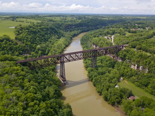 Aerial view of a rusted railway bridge spanning a muddy river, surrounded by lush green trees and rolling hills.