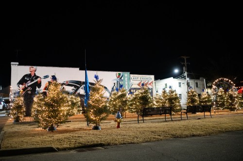 A festive scene with decorated Christmas trees and lights at night, featuring a mural and benches in the background.
