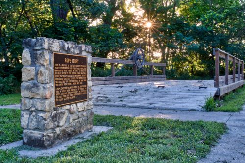 A stone plaque beside a wooden bridge in a wooded area, with sunlight filtering through the trees in the background.