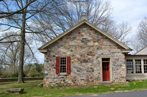 A stone house with red shutters and a red door, surrounded by trees and grass under a cloudy sky.