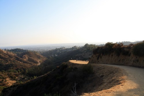 A winding path through a hilly landscape, overlooking a distant city under a clear sky.
