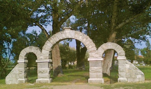 Stone archway with inscriptions, surrounded by trees in a grassy area, leading to a memorial or park.