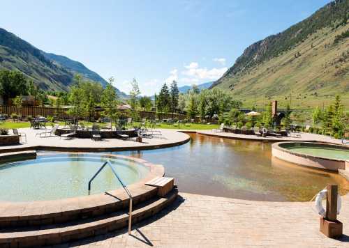 A scenic outdoor spa with hot tubs and lounge chairs, surrounded by mountains and greenery under a clear blue sky.