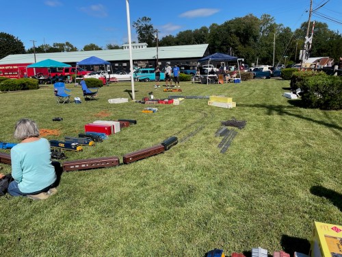A grassy area with model trains laid out, people sitting nearby, and tents in the background under a clear blue sky.
