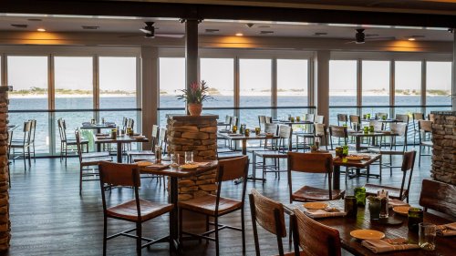 A bright restaurant interior with wooden tables and chairs, large windows overlooking a waterfront view.