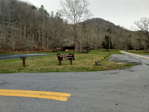 A grassy area with signs near a road, surrounded by bare trees and mountains in the background on a cloudy day.