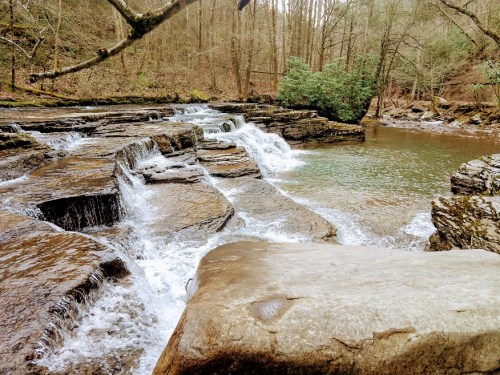 A serene waterfall cascades over rocky ledges into a calm pool, surrounded by trees and natural greenery.