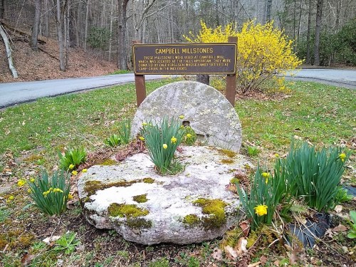 A historic millstone surrounded by flowers and grass, with a sign reading "Campbell Millstones" in a wooded area.