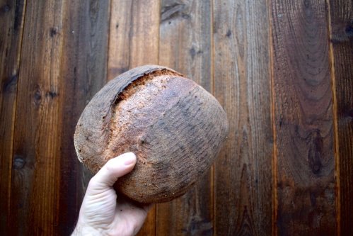 A hand holding a round, rustic loaf of bread against a wooden background.