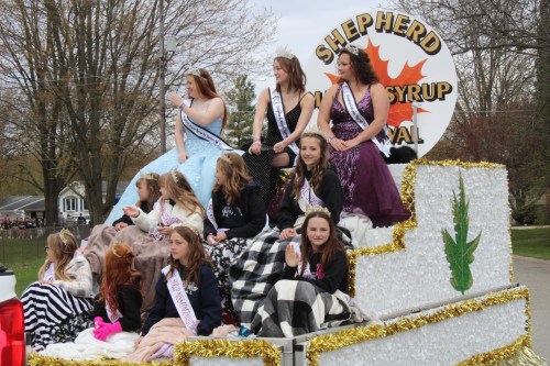 A parade float featuring young women in formal dresses, some wrapped in blankets, celebrating a syrup festival.