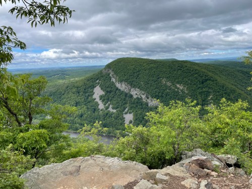 A scenic view of a green mountain surrounded by trees, with a river winding through the valley below under a cloudy sky.