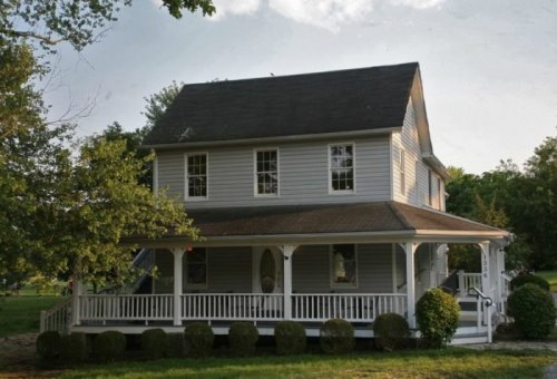 A two-story gray house with a porch, surrounded by greenery and trees under a cloudy sky.