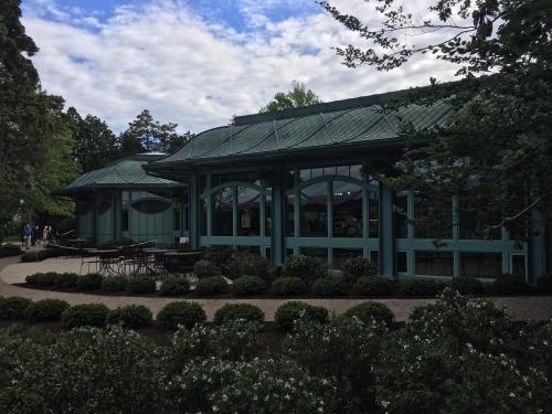 A green building with large windows surrounded by trees and shrubs, featuring outdoor seating areas.