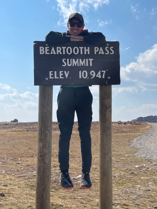 Person in black attire playfully hanging on a sign that reads "Beartooth Pass Summit Elev. 10,947" against a scenic backdrop.