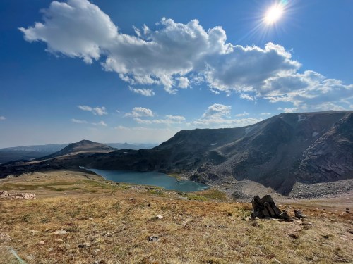 A scenic view of a mountain landscape with a lake, grassy terrain, and a bright blue sky with scattered clouds.