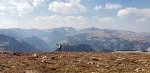 A person stands with arms outstretched on a rocky mountain landscape, surrounded by distant peaks and a blue sky.