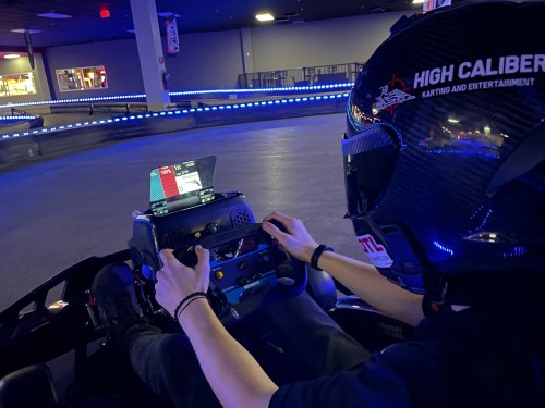 A person in a racing helmet adjusts controls in a go-kart on a brightly lit indoor track.