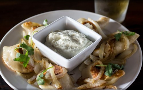A plate of dumplings topped with sautéed onions, served with a creamy dip and garnished with cilantro.